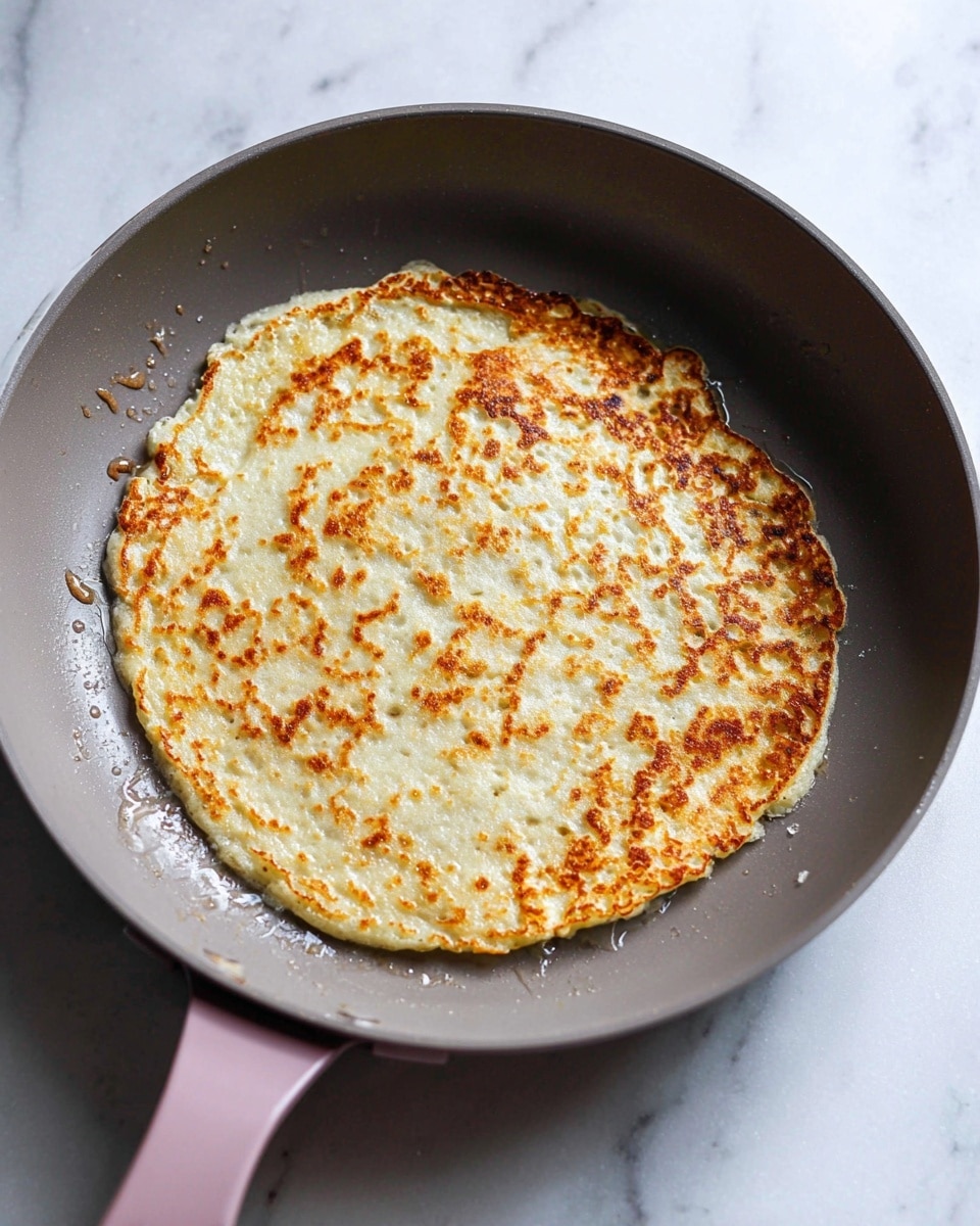 A single round pancake is shown cooking in a gray pan with a pink handle on a white marbled surface. The pancake has a golden-brown, slightly crispy outer layer with an uneven texture, showing small bubbles and a few darker spots where it is more cooked. The pancake's center is a lighter beige color, appearing soft and thick. There are small bits of batter that have spread out near the edges, giving the pancake a natural, homemade look. The pan has some oil droplets around the pancake, adding a slight shine to the surface. Photo taken with an iphone --ar 4:5 --v 7