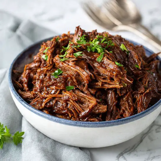A close-up image of a bowl filled with shredded meat covered in a thick, rich, dark brown sauce. The bowl is white with a blue outer edge, and it is placed on a surface with a white marbled texture. On top of the meat, there are a few sprigs of fresh green herbs for garnish. In the background, there is a soft, light gray cloth with slightly blurred silver forks resting on it. photo taken with an iphone --ar 4:5 --v 7