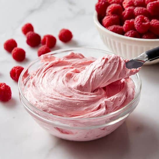 A clear glass bowl filled with smooth, thick, pink frosting that has soft swirls on the surface. A black-handled spatula lies on top, holding a generous layer of the same pink frosting, showing a creamy texture. Behind the bowl, there is a white bowl full of fresh red raspberries, with a few loose raspberries placed on a white marbled surface. The scene is bright and clean with a soft focus on the background, emphasizing the rich pink frosting in the foreground photo taken with an iphone --ar 4:5 --v 7