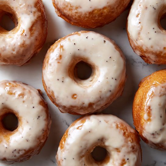 The image shows a close-up of about ten glazed doughnuts arranged closely together on a white marbled surface. Each doughnut has a rough, slightly uneven texture with a thick shiny glaze layer that looks white and smooth but with some translucence showing the golden brown dough beneath. The doughnuts have a ring shape with a well-defined hole in the middle and some darker spots where the baked dough peeks through. The glaze is unevenly applied, pooling in some areas and lighter in others, giving each doughnut a slightly different appearance. The overall color palette includes warm golden browns, creamy whites, and small dark specks scattered throughout the dough. photo taken with an iphone --ar 4:5 --v 7