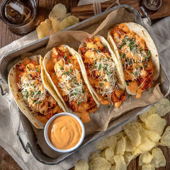 The image shows a metal tray lined with parchment paper holding five soft white tortillas folded in half. Each tortilla is filled with a layer of melted orange cheese sauce at the base, topped with pieces of cooked reddish meat, and finished with plenty of light-colored melted shredded cheese and small green herb sprigs. The tray is placed on a wooden surface with scattered pale yellow potato chips around it. A small round white bowl of creamy orange dipping sauce is placed inside the tray near the chips. In the background, a glass with a dark drink and a metal bottle cap on a beige cloth napkin are visible. photo taken with an iphone --ar 4:5 --v 7