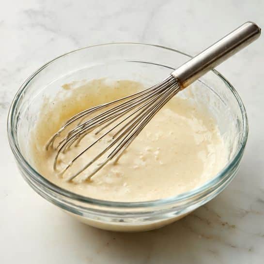 A clear glass bowl filled with a light beige, creamy batter that has a smooth but slightly thick texture, with some small lumps visible. A metal whisk with a silver handle is placed inside the bowl, partially covered in the batter. The bowl is set on a white marbled surface. photo taken with an iphone --ar 4:5 --v 7