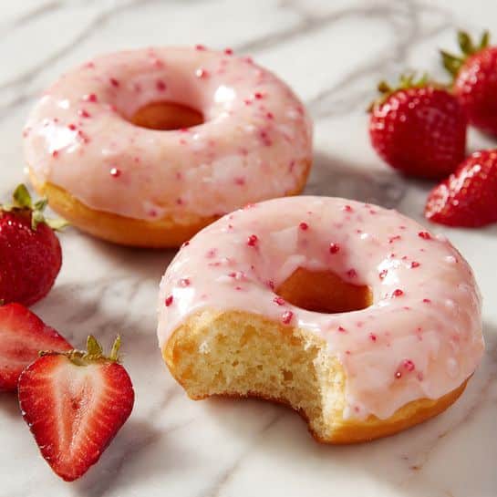 Two light, fluffy donuts are shown on a white marbled surface. Each donut has a smooth, shiny pink glaze with small red specks evenly spread. The donut in the front has a bite taken from it, revealing its soft, pale yellow inside. Around the donuts, there are fresh halved strawberries with bright red flesh and green leaves. The image looks fresh and colorful, with soft natural light. Photo taken with an iphone --ar 4:5 --v 7