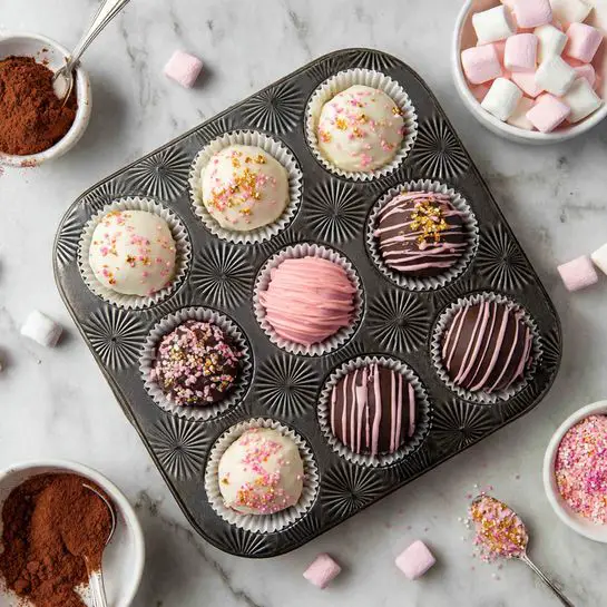 The image shows six round cupcakes in a dark metal muffin tray with star patterns, each cupcake in white paper liners. The cupcakes have three different tops: smooth white chocolate with pink and gold sprinkles and drizzle; smooth pink chocolate with dark chocolate drizzle and pink and gold sprinkles; and smooth dark chocolate with light pink drizzle. Around the tray, on a white marbled surface, there are small pink marshmallows, loose pink, gold, and white sprinkles. To the left, a white bowl with cocoa powder and a silver spoon inside it is visible, while two white bowls with pink and gold sprinkles sit to the right and bottom. One cupcake is open, showing chocolate powder and pink marshmallows inside its white chocolate shell. Photo taken with an iphone --ar 4:5 --v 7