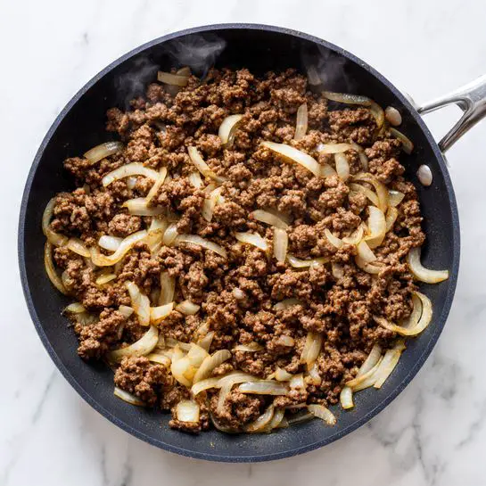 A large black frying pan filled with cooked ground meat mixed with small pieces of soft, translucent cooked onions. The meat is brown with a crumbly texture, and the onions are light yellow with a glossy surface. The pan is on a white marbled surface, showing some steam rising gently. The image is taken from above, focusing on the even spread of meat and onions inside the pan. photo taken with an iphone --ar 4:5 --v 7
