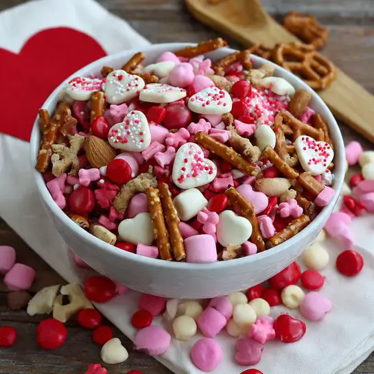 A white bowl filled with a colorful mix of pink yogurt-covered pretzels, red and pink candy-coated chocolates, white and pink marshmallows, peanuts, raisins, and small animal-shaped crackers. On top, heart-shaped white chocolate pieces with red sprinkles add a decorative touch. The bowl is placed on a wooden surface next to a wooden paddle. A white cloth with a large red heart and scattered pink and red candies lies below the bowl, with some candies spilling onto the wooden surface. photo taken with an iphone --ar 4:5 --v 7