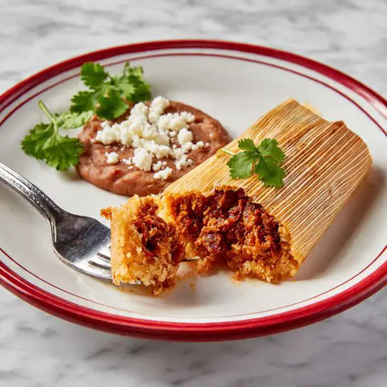 A white plate with a red rim holds a tamale partially wrapped in a light brown corn husk lying flat across the plate. The tamale is golden brown, with a soft, crumbly texture on the outside, and a rich dark reddish-brown filling visible where a piece has been broken off and speared on a silver fork resting on the plate. Behind the tamale is a round dollop of brown refried beans topped with small white crumbles, arranged neatly on the plate. Around the plate, some green cilantro leaves add a fresh touch, all set on a white marbled surface. photo taken with an iphone --ar 4:5 --v 7