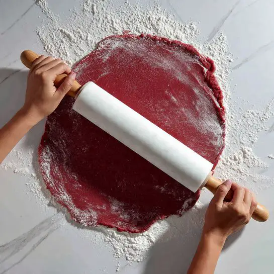 A close-up image shows a pair of woman's hands holding a white marble rolling pin with wooden handles, rolling out a smooth, deep red dough that is dusted with white flour on a white marbled surface. The dough is evenly rolled into a large, thin oval shape with flour patches scattered across it. The scene captures the texture of the dough and the gentle pressure from the rolling pin, highlighting the contrast between the vivid red dough and the white flour and surface. photo taken with an iphone --ar 4:5 --v 7