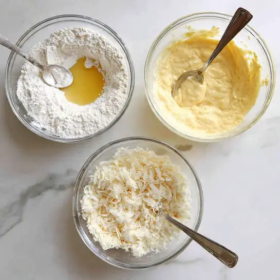 The image shows three clear glass bowls on a white marbled surface, each with different stages of batter preparation. In the first bowl, white flour surrounds a pale yellow liquid that is being poured in, with a spoon resting inside the bowl at the top right. The second bowl contains a smooth, creamy light yellow batter with a spoon partially dipped into it on the right side. The third bowl shows the same light yellow batter with white shredded pieces on top, with the spoon resting on the right, partially mixed in. photo taken with an iphone --ar 4:5 --v 7