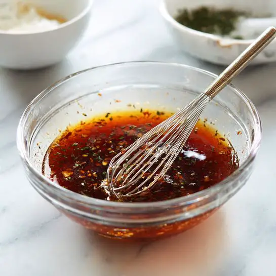 A clear glass bowl holds a dark reddish-brown liquid mixture with visible small herb flakes floating throughout, creating a textured and rich spicy appearance. A silver metal whisk rests inside the bowl, its wires partially submerged in the liquid, with splatters of the mixture seen around the inside edges of the bowl. The bowl sits on a white marbled surface, and there is a blurred white bowl with some light ingredients in the background. photo taken with an iphone --ar 4:5 --v 7