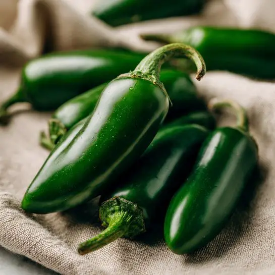 The image shows a close-up of several bright green jalapeño peppers lying together on a beige fabric with a soft texture. The peppers have a smooth, shiny skin with a few highlights showing their freshness. In the background, more jalapeños blur softly, adding depth to the picture. The overall scene is simple, focusing only on the peppers with a white marbled textured surface underneath. photo taken with an iphone --ar 4:5 --v 7