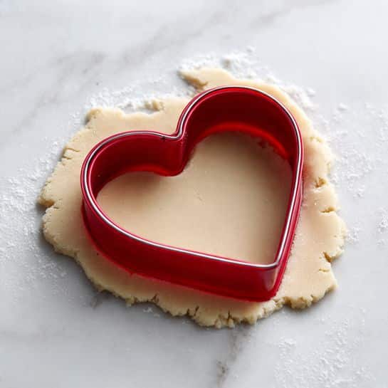 A close-up view shows a red heart-shaped cookie cutter pressed into a flat layer of pale dough. The dough inside the cookie cutter is smooth and fills the entire heart shape, while the surrounding dough is rolled out thinly with a slightly uneven surface. The setup rests on a white marbled surface, providing a clean and bright background. The image captures the moment before the heart-shaped dough is removed from the larger sheet photo taken with an iphone --ar 4:5 --v 7