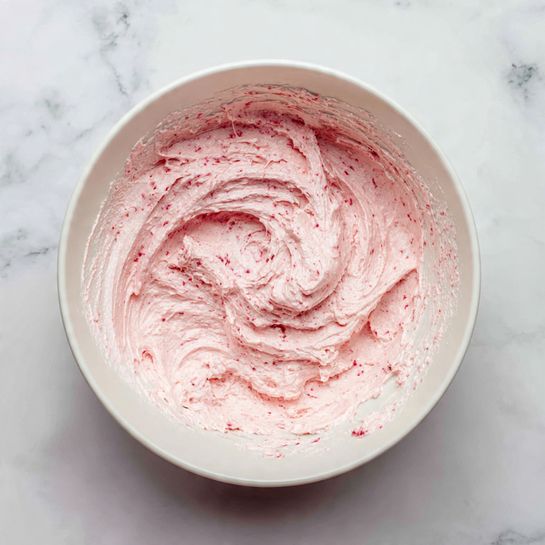 Inside a large white bowl, there is a thick, creamy pink mixture with a slightly fluffy texture. The mixture is swirled in the bowl, showing some small darker pink bits mixed throughout. The edges of the bowl have remnants of the mixture sticking to the sides. The bowl is set on a white marbled textured surface. photo taken with an iphone --ar 4:5 --v 7