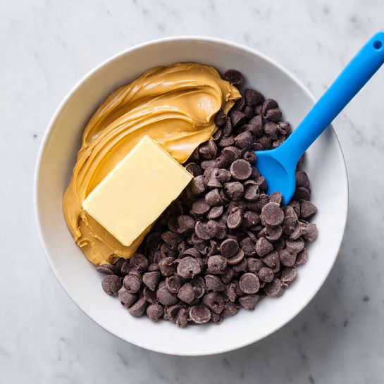 A white bowl contains three main layers of ingredients: a large portion of dark brown chocolate chips covering about half the bowl, a smooth layer of light brown peanut butter spread next to the chocolate, and a stick of pale yellow butter resting on top of the chocolate chips near the peanut butter. A blue spatula is partly dipped into the peanut butter. The bowl is placed on a white marbled surface. photo taken with an iphone --ar 4:5 --v 7