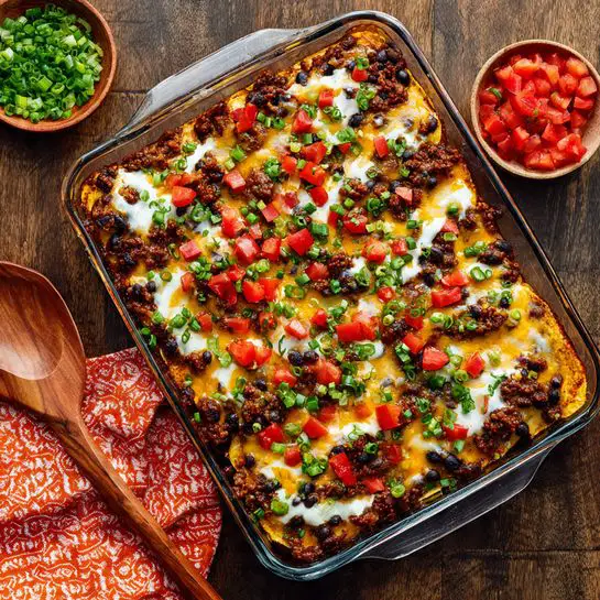 A rectangular clear glass baking dish filled with three layers of food: the bottom layer is pale yellow soft tortillas, the middle layer is a mix of browned ground meat with black beans and small bits of red and green vegetables, and the top layer is melted cheese in white and yellow colors covering the whole dish. On top, small chopped bright red tomatoes and chopped green onions are scattered mostly in the middle. The dish sits on a dark wooden surface with some chopped tomatoes and green onions on a wooden spoon to the bottom left and an orange cloth with white patterns on the right. Photo taken with an iphone --ar 4:5 --v 7