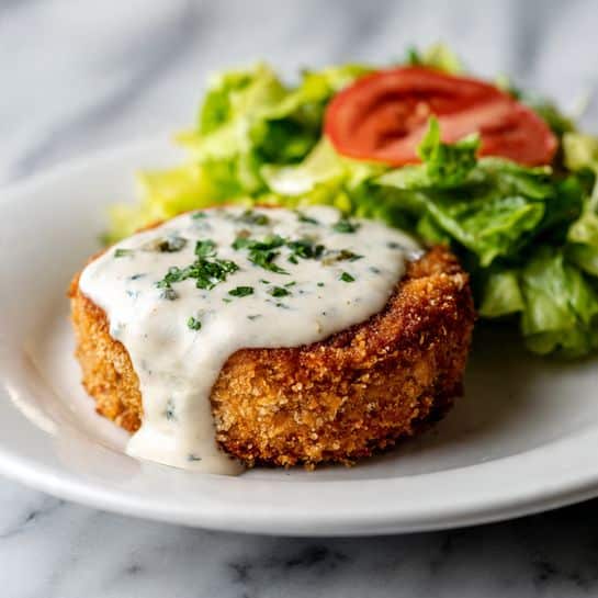 A white plate on a white marbled surface holds a golden brown round fried patty with a crispy texture. On top of the patty is a smooth, creamy white sauce drizzled, with small bits of green herbs sprinkled over it. Behind the patty is a side salad with bright green lettuce leaves and a few red tomato slices, adding color contrast. The whole scene is softly lit, showing the details of the crispy patty and fresh salad clearly. Photo taken with an iphone --ar 4:5 --v 7