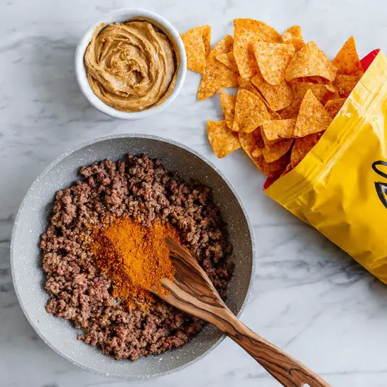 A light gray pan holds cooked ground meat covering the whole surface; in the center, there is a mound of bright orange spice powder. A wooden spatula with visible grain rests on top of the meat, partially submerged. Above the pan is an open yellow bag spilling orange seasoned chips onto a white marbled surface. To the left of the pan, there is a small white bowl filled with dense, chunky peanut butter. Photo taken with an iphone --ar 4:5 --v 7