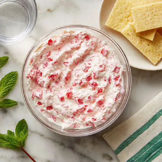 Stuffed Wonton Bites with Strawberry Cream Filling Recipe 4 A clear glass mixing bowl sits on a white marbled surface. Inside the bowl is a creamy white mixture with small chunks of bright red, creating a textured look. The mixture is spread slightly around the sides of the bowl, showing its smooth and thick consistency. Nearby on the surface, a white plate holds several pale yellow rectangular sheets with a slightly powdery texture. There are a few green mint leaves adding a fresh touch. A small clear glass bowl filled with water is also visible. A cloth with green and white stripes lies folded on the side. photo taken with an iphone --ar 4:5 --v 7