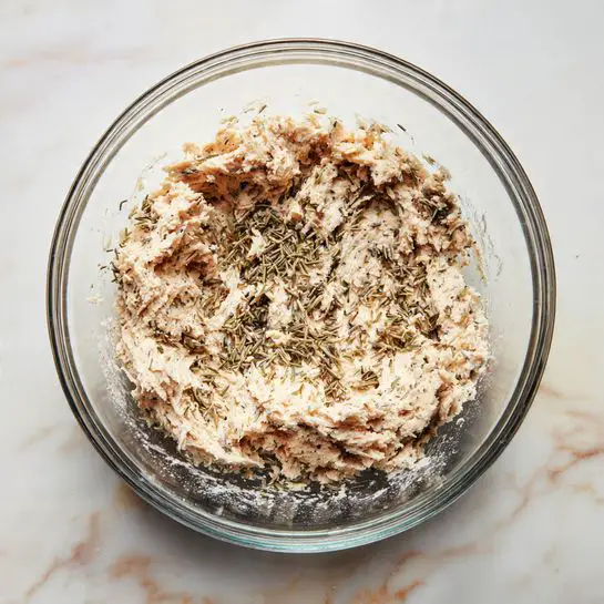 The image shows a clear glass bowl placed on a white marbled surface, containing a single layer of a pale, creamy mixture with small bits of herbs and possibly shredded meat evenly spread. The texture looks soft and slightly chunky, and the mixture is roughly centered inside the bowl with some residue around the edges. Photo taken with an iphone --ar 4:5 --v 7