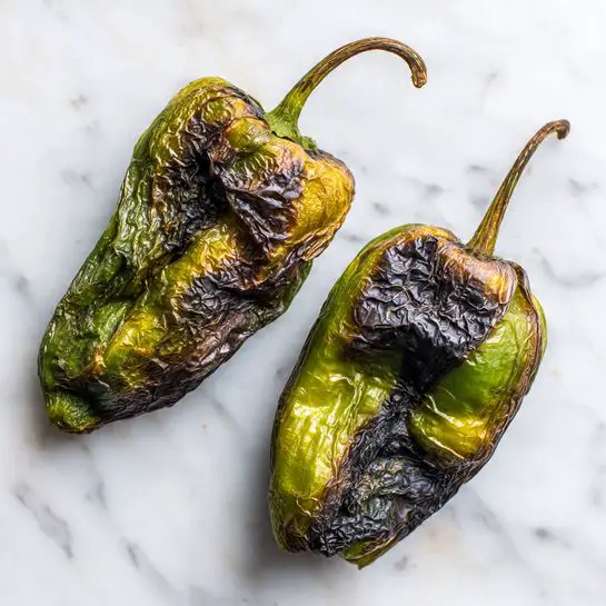 Two roasted green peppers are shown lying side by side on a white marbled surface. Both peppers have dark, wrinkled skin with patches of brown and black from roasting, showing a rough, charred texture. The peppers are slightly shriveled with uneven, dimpled surfaces and visible stems attached at one end. The overall colors range from deep green to burnt black and brown, highlighting their roasted state. Photo taken with an iphone --ar 4:5 --v 7