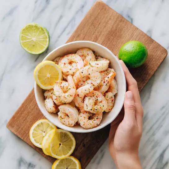 A white bowl filled with many cooked shrimp with a light pink and white color, placed on a wooden board. Nearby, there are two lemon wedges with bright yellow color and a whole green lime. A woman's hand is holding the top edge of the bowl. The background is a white marbled surface. photo taken with an iphone --ar 4:5 --v 7