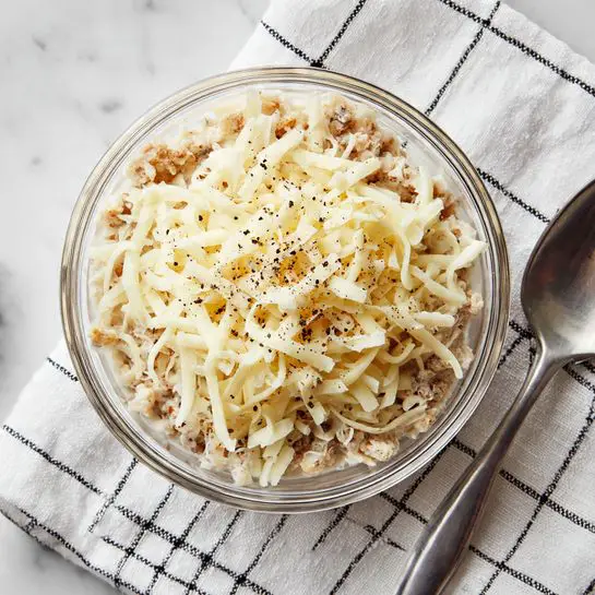 A clear glass bowl holds a layered mixture on a white marbled surface. The bottom layer is chunky and light brown, topped by a generous layer of shredded pale yellow cheese with some black pepper sprinkled over it. The bowl sits on a white cloth with a black grid pattern, and a silver spoon lies next to it on the right side. Photo taken with an iphone --ar 4:5 --v 7