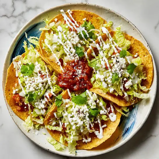 Three folded golden corn tortillas with a slightly crispy texture are arranged in a circle on a white plate with blue swirls. Beneath and around the tortillas is a bed of shredded light green lettuce. The tortillas are topped with scattered fresh cilantro leaves, white creamy sauce drizzled in thick lines, a dollop of dark red chunky salsa in the center, and a light sprinkling of white crumbly cheese. The plate rests on a white marbled surface. photo taken with an iphone --ar 4:5 --v 7