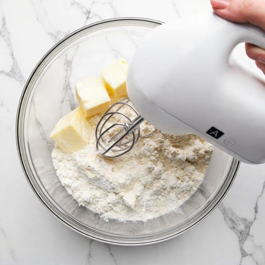 A close-up image shows a woman's hand holding a white electric mixer inside a clear glass bowl. The bowl contains a few pieces of pale yellow butter and white flour powder. The background surface has a white marbled texture. The mixer is white with a digital display showing the letter