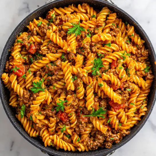 A close-up view of a pan filled with rotini pasta cooked with ground beef and small pieces of tomato, all mixed well in a rich reddish-orange sauce. The pasta spirals are bright orange, coated evenly in sauce, and scattered with bits of brownish ground beef and small red tomato chunks. The dish is sprinkled with fresh green parsley leaves, adding a pop of color on top. The pan is black with a textured edge, sitting on a white marbled surface. photo taken with an iphone --ar 4:5 --v 7