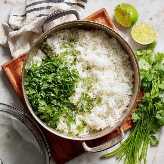 A pot filled with fluffy white rice makes up the bottom layer, topped with a bright green pile of chopped cilantro on one side. The pot has a shiny metal handle and is sitting on a wooden square trivet. Nearby, on a white marbled surface, there is a halved lime showing its inside and a bunch of fresh cilantro with long stems and vibrant leaves. A clear glass lid is leaning against the pot, and a cloth with black and white stripes is slightly visible in the corner. The light is soft, creating a fresh and clean feeling. photo taken with an iphone --ar 4:5 --v 7