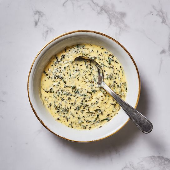 A white bowl with an uneven gold rim contains a creamy, thick yellow sauce mixed with small green herb bits spread evenly throughout. A spoon with a silver bowl and a black handle rests inside the bowl, partially coated with the sauce. The bowl is placed on a white marbled surface. photo taken with an iphone --ar 4:5 --v 7