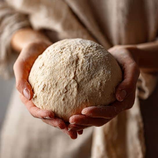 A woman's hand gently holds a smooth, round ball of dough with a light beige color and a soft texture. The dough looks fresh and slightly dusted with flour, showing small natural creases on the surface. The background is blurred with soft neutral tones, making the dough the main focus. The photo taken with an iphone --ar 4:5 --v 7