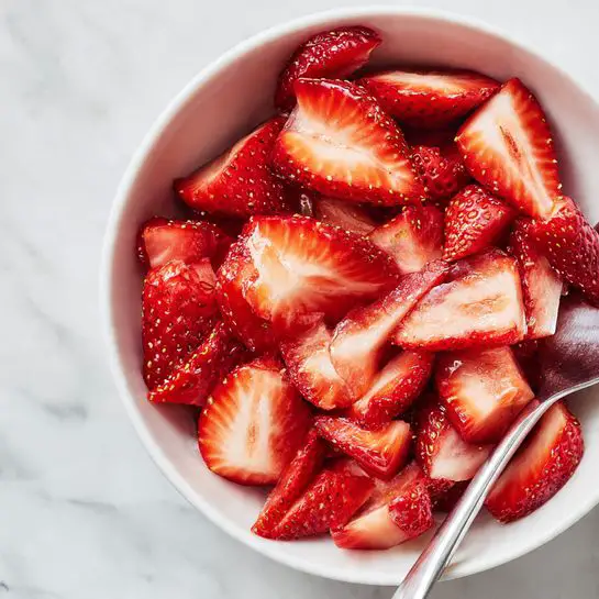 The image shows a close-up view of many sliced strawberries inside a white bowl. The strawberries have a bright red outer layer with visible yellow seeds, and the inside is a lighter, almost pale pink color with a soft texture. The slices vary in size and are tightly packed, showing their juicy and fresh look. Part of a silver spoon is visible on the right side, resting among the strawberry slices. The background is a white marbled surface, giving a clean and fresh feel to the image. photo taken with an iphone --ar 4:5 --v 7
