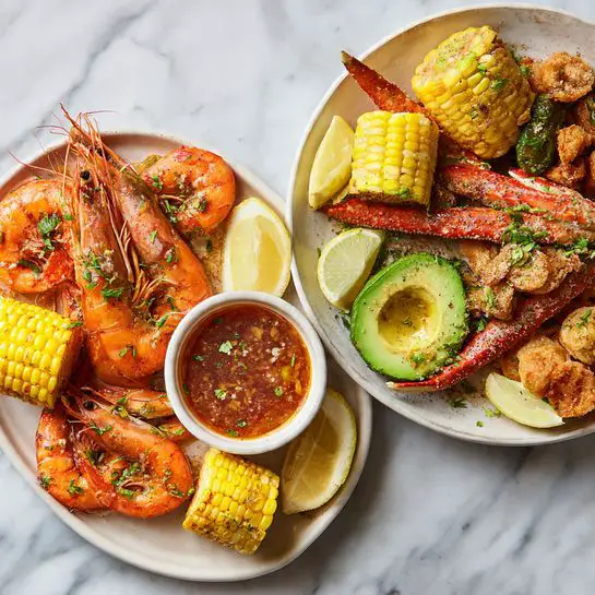 The image shows two seafood dishes side by side on a white marbled texture. The left dish is arranged on a white plate with several cooked orange shrimp topped with green herbs, two pieces of bright yellow corn on the cob, and a bowl of reddish-brown sauce with visible pieces inside. The shrimp surround the bowl, and lemon halves are placed around the plate. The right dish is served on a white plate with a mix of seafood including cooked red crab claws, orange shrimp, and pieces of shellfish. A small bowl with reddish sauce sits in the center, surrounded by yellow corn on the cob, lemon wedges, a green avocado half, and some fried food. Both dishes have a rustic and fresh appearance. photo taken with an iphone --ar 4:5 --v 7