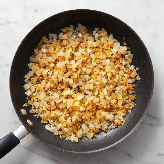 A black frying pan filled with a single layer of small, cooked onion pieces that are golden brown with some darker crisp edges, showing a mix of soft and slightly crispy textures. The pan's handle extends out from the left side, resting on a white marbled surface that adds a clean and bright contrast to the warm tones of the onions. The onions cover the bottom of the pan evenly, with a few small bits scattered around the edges. photo taken with an iphone --ar 4:5 --v 7