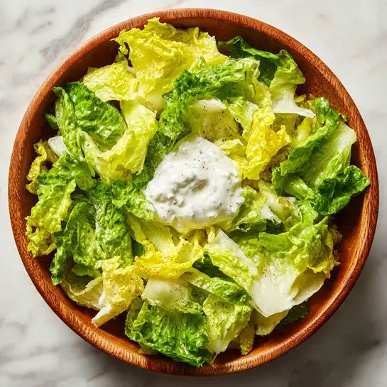 A wooden bowl filled with fresh, chopped lettuce leaves in shades of bright green and yellow, topped with a dollop of creamy white dressing sitting in the center. The lettuce has a crisp texture with some darker green leaves mixed with lighter ones, and the dressing looks smooth and thick, starting to blend slightly into the greens. The bowl is placed on a white marbled surface. Photo taken with an iphone --ar 4:5 --v 7