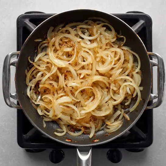 A black frying pan on a stove holds a single layer of finely sliced onions cooking until they are golden brown and soft, showing some light caramel color with slightly translucent edges. The pan has silver handles on each side and the stove beneath it is dark with silver accents. The background surface is a white marbled texture. photo taken with an iphone --ar 4:5 --v 7