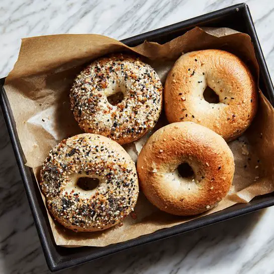 Four bagels on white parchment paper on a round grey tray, two of the bagels topped with mixed seeds like sesame and poppy, the other two plain with a golden brown crust, the bagels have a slightly shiny texture and soft fluffy look, next to the tray is a small white bowl with a dollop of cream cheese and a clear glass cup with dark coffee, all set on a white marbled surface, a silver knife with some cream cheese on it is resting near the tray, a striped cloth partially in the bottom left corner, photo taken with an iphone --ar 4:5 --v 7