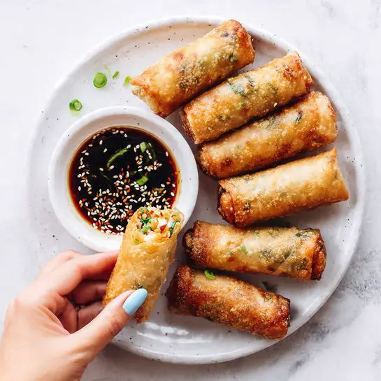 On a white plate with a white marbled surface underneath, five golden brown spring rolls with slightly shiny, crispy skins are lined up. Each spring roll has bits of green herbs visible on the outside, and small green onion pieces are sprinkled both on them and on the plate. A white ceramic dipping bowl filled with dark soy sauce sprinkled with white sesame seeds sits in the center. A woman's hand with light blue painted nails is holding one spring roll, dipping it halfway into the sauce. The scene is bright and clear, showing textures of the spring rolls and the sauce. photo taken with an iphone --ar 4:5 --v 7