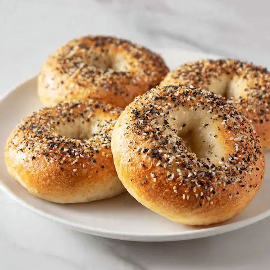 The image shows four round bagels with a hole in the middle, placed close together on a white plate. Each bagel has a shiny, golden-brown crust covered with white sesame seeds and small black poppy seeds evenly spread on top. The bagels have a slightly rough texture with a few visible cracks, showing a soft inside under the crust. The background is a white marble surface. photo taken with an iphone --ar 4:5 --v 7