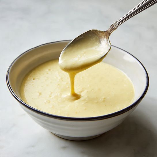 A white bowl with a dark rim is filled with smooth, creamy batter that is light yellow in color and has a few small bubbles on the surface. A spoon with a silver head and a black handle is held above the bowl, dripping some of the batter back into it, showing its thick but flowing texture. The bowl sits on a white marbled surface that adds a clean and simple background to the image. Photo taken with an iphone --ar 4:5 --v 7