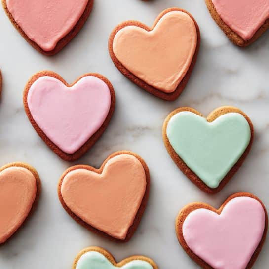 The image shows stacks of heart-shaped cookies with three visible layers. Each cookie has a red base layer with a slightly rough texture, topped by a smooth, colorful icing layer in pastel shades of pink, yellow, and light green. The top cookie in the front stack has pink icing with a small bite taken from its right curve, revealing the red base underneath. The cookies are arranged on a white marbled surface with a pink and white striped cloth in the background. The lighting is soft and natural, emphasizing the bright colors and textures of the cookies. Photo taken with an iphone --ar 4:5 --v 7