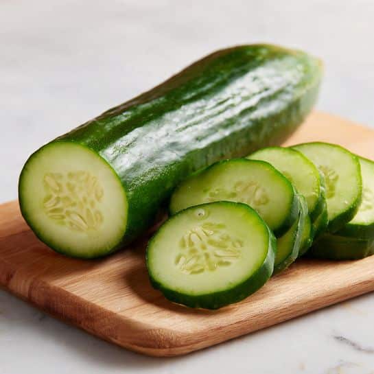 A white bowl filled with a bottom layer of white rice sprinkled with chopped green onions, topped with several golden brown crispy fried slices layered closely side by side, each piece showing a white interior and a crunchy textured crust. A thick dark brown sauce is drizzled over the fried slices, garnished with white sesame seeds and more green onions. To the side, there is a neat layer of thinly sliced green cucumbers arranged in a row. The bowl sits on a white marbled surface in soft daylight. Photo taken with an iphone --ar 4:5 --v 7