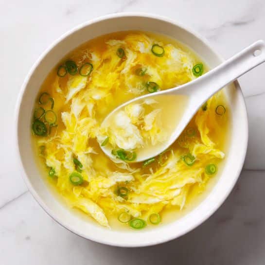 A white bowl filled with soft, yellow egg drop soup with visible silky ribbons of cooked egg floating in a clear, light yellow broth. Thin, fresh green slices of scallion are scattered gently on the surface, adding small bright green accents. A white ceramic spoon rests inside the bowl, lifting a portion of the soup with egg ribbons and scallions clearly visible on it. The bowl is placed on a white marbled surface. Photo taken with an iphone --ar 4:5 --v 7
