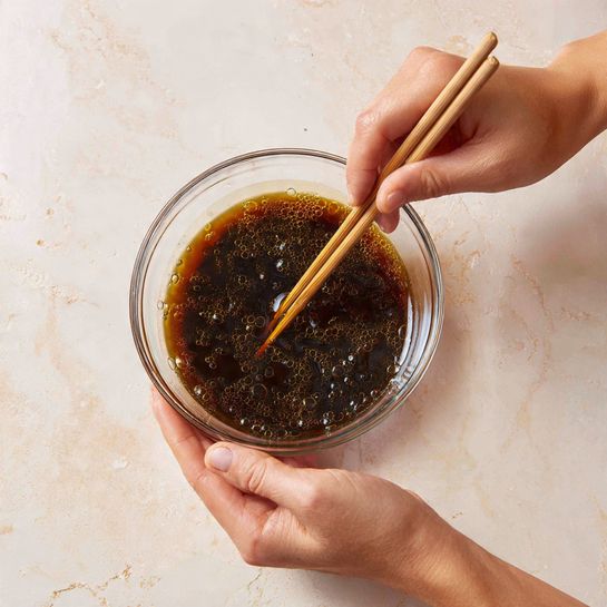 A clear glass bowl is held by a woman's hand on the right side, containing a thick, dark brown liquid being gently stirred with light brown wooden chopsticks held by a woman's hand on the left. The liquid has a glossy texture with small bubbles on the edges, and the background is a white marbled surface photo taken with an iphone --ar 4:5 --v 7