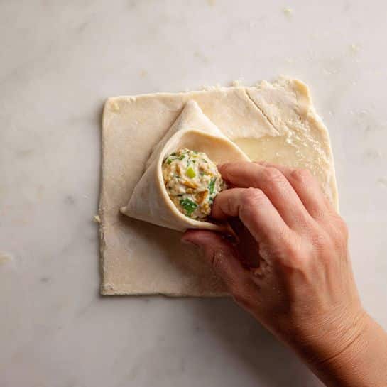 A woman’s hand is folding a square piece of thin, light beige dough around a small round ball of creamy filling that has visible green leafy herbs and small bits of light brown pieces. The dough rests flat on a white marbled surface and has an area brushed with a shiny wet substance along one edge. The hand is pinching the dough to start wrapping the filling, showing the texture and gentle folds of the dough near the filling photo taken with an iphone --ar 4:5 --v 7