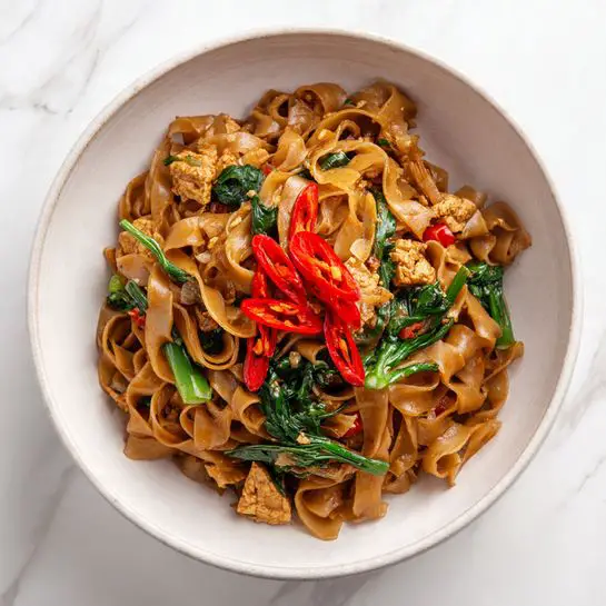 A bowl filled with a colorful stir-fried dish showing about three layers: the bottom layer is wide, flat, slightly shiny brown noodles tangled together; scattered throughout the noodles are small, light tan chunks of tofu; mixed in are bright green vegetable leaves and herbs adding fresh contrast, with pieces of sliced red chili peppers spread on top for a touch of color and heat; the bowl itself is white with a matte texture, placed on a white marbled surface. photo taken with an iphone --ar 4:5 --v 7