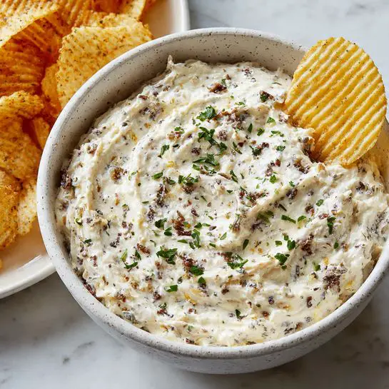 A white speckled bowl filled with a thick, creamy dip that has small brown bits mixed throughout, topped with small green parsley pieces scattered on the surface. A ridged potato chip is dipped into the creamy mixture, standing upright on the right side of the bowl. In the background, there is a white plate with more ridged potato chips, all placed on a white marbled surface. photo taken with an iphone --ar 4:5 --v 7
