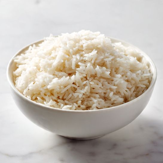 A white ceramic bowl filled with a round mound of fluffy white rice. The rice grains look soft and separate, with a slightly shiny texture, filling the bowl nearly to the top. The bowl sits on a white marbled surface with soft natural light that highlights the rice's texture and the bowl's smooth finish. photo taken with an iphone --ar 4:5 --v 7