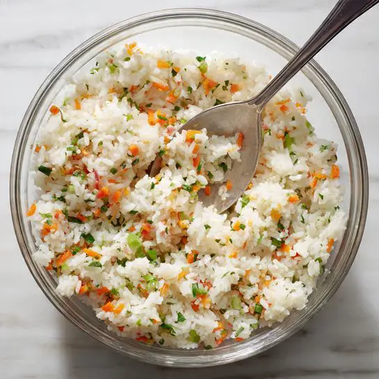 A clear glass bowl filled with a mixture of cooked white rice, finely chopped small orange carrot pieces, and tiny bits of green vegetable, all mixed together. A metal spoon is partially visible inside the bowl, scooping the rice mixture. The bowl is placed on a white marbled surface. The colors are soft and natural, with the white rice as the main base, dotted with the bright orange and green bits evenly spread throughout. Photo taken with an iphone --ar 4:5 --v 7